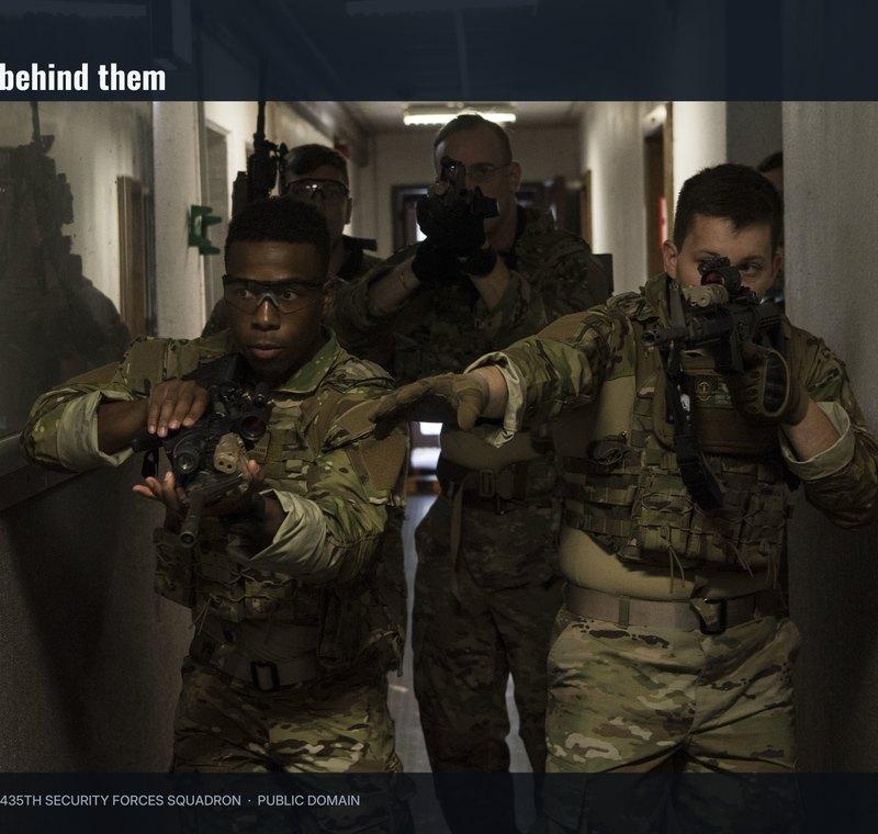 A team of airmen with carbines advancing down a narrow hotel style corridor during close quarters battle training
