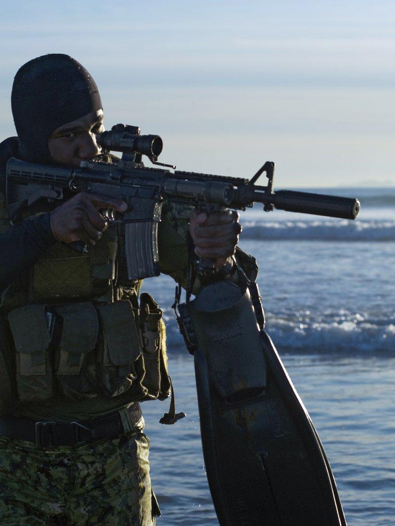 Navy SEAL in a neoprene hood standing in the surf with fins in hand, aiming a suppressed short carbine with a fixed front sight and red dot