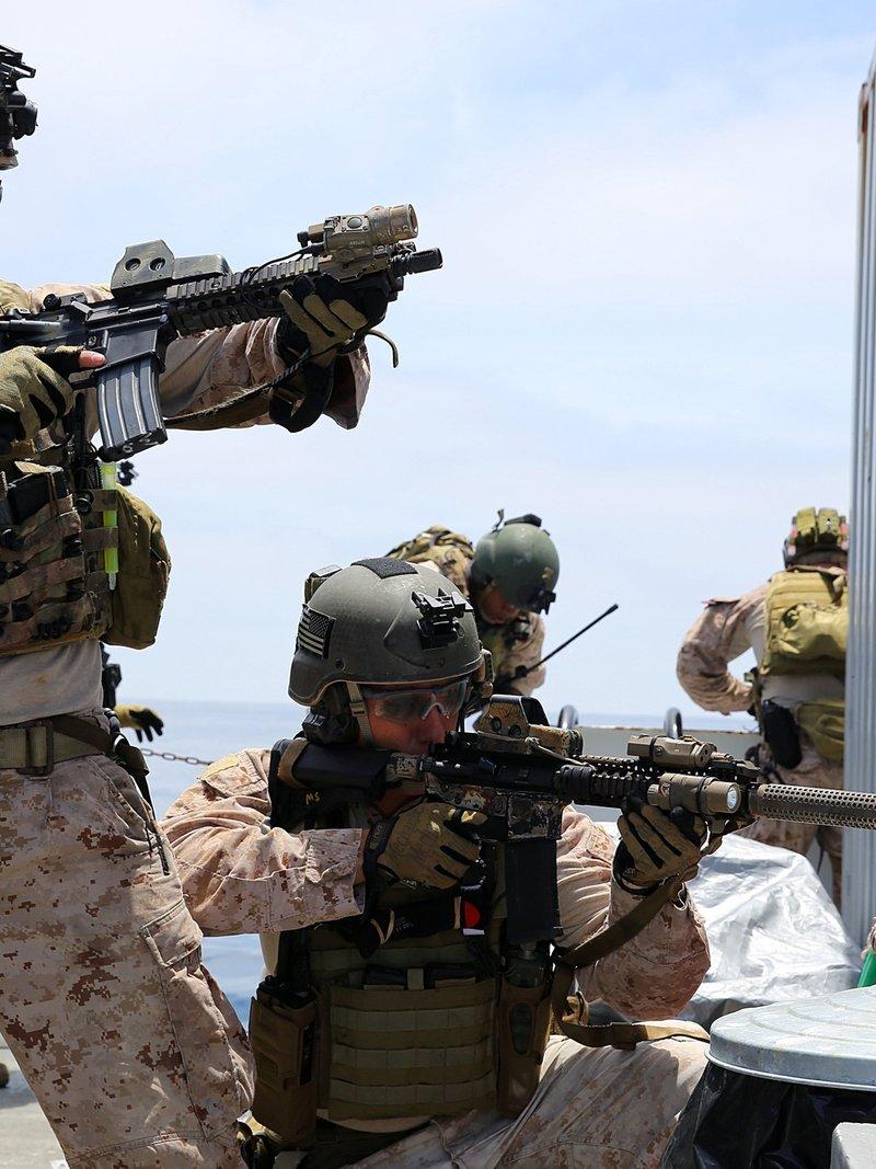 Two Marines on a ship deck in desert camouflage aiming short barreled rifles with black quad rails, lasers, a weapon light and a suppressor