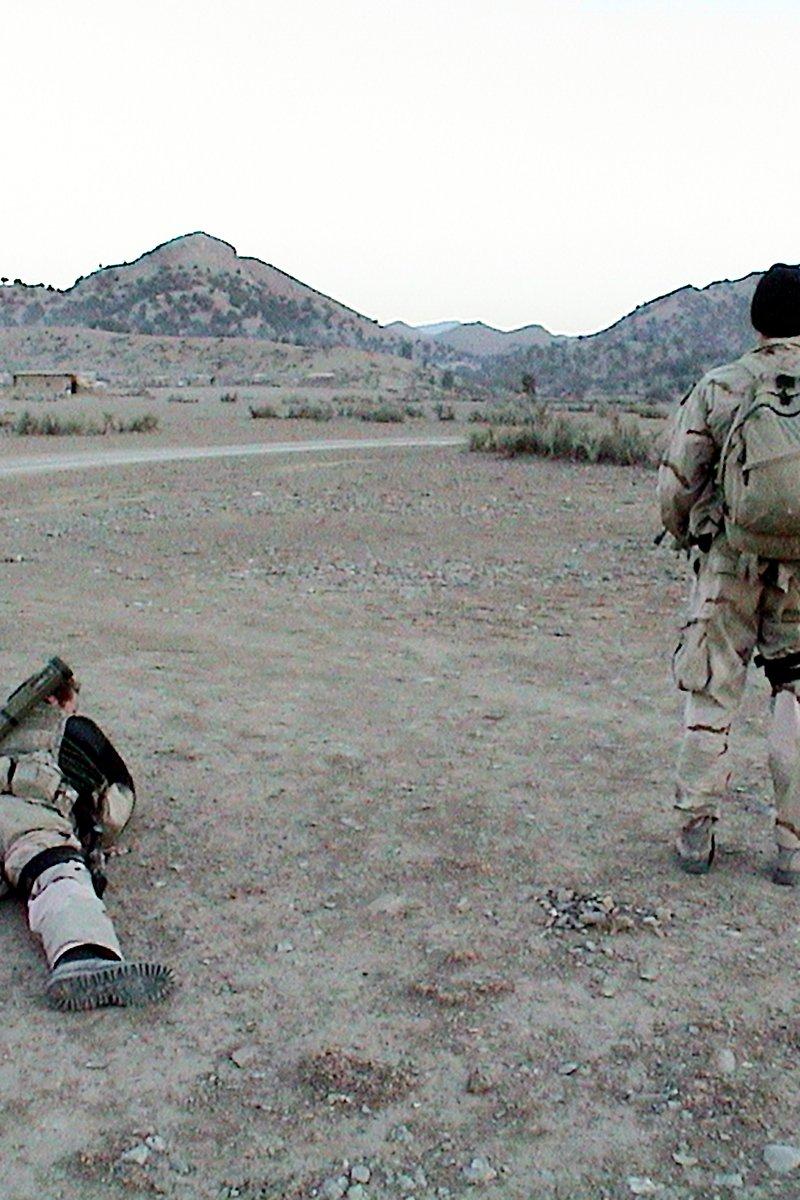 Two U.S. Navy SEALs in desert uniforms on open ground in eastern Afghanistan, one prone with a launcher on his back, facing low mountains and a few buildings