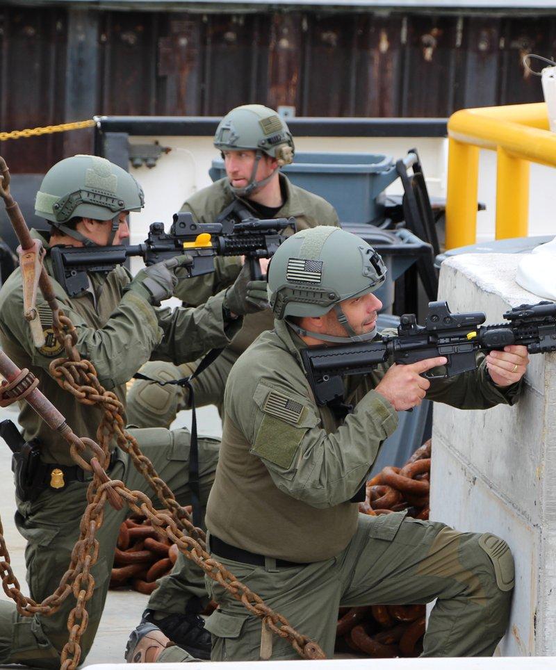 Detroit FBI SWAT members in olive uniforms and helmets take cover behind a concrete block on a pier, two of them aiming black carbines with quad rails and EOTech sights