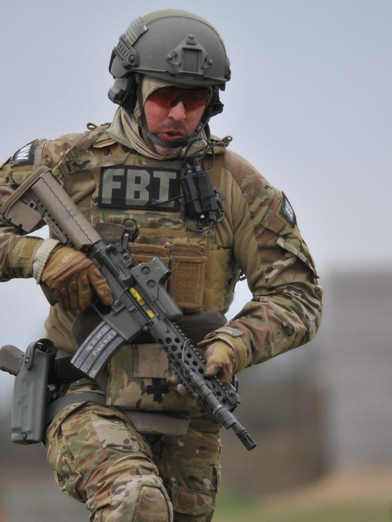 A Milwaukee FBI SWAT member in Multicam and an FBI marked plate carrier moves across a training range with a short carbine fitted with a round hole free float rail, an EOTech sight and a weapon light