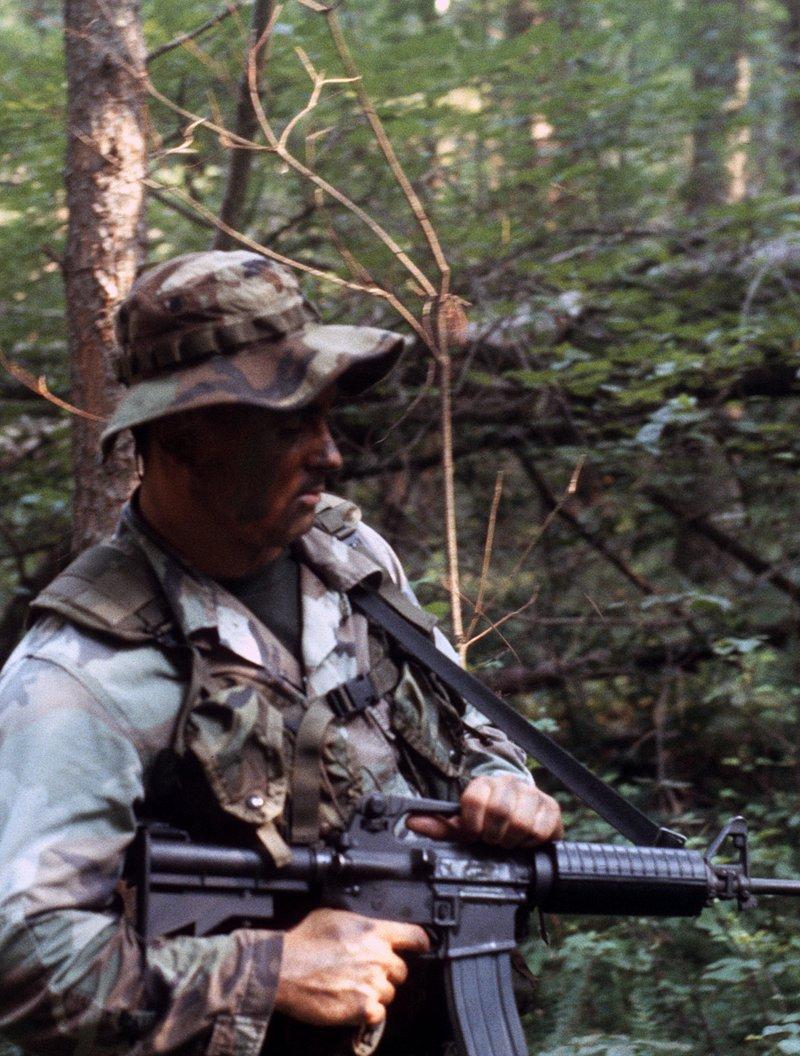 A Scout Sniper Instructor School student in woodland camouflage and a boonie hat carries an M16A2 carbine with round handguards on patrol in the woods at Quantico in 1987