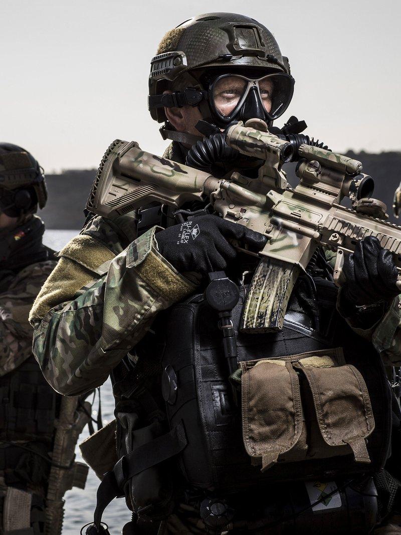 Dutch commando diver in a mask and rebreather holding a camouflage painted HK416 D10RS with a quad rail, magnified optic and light, two divers behind him at the water's edge