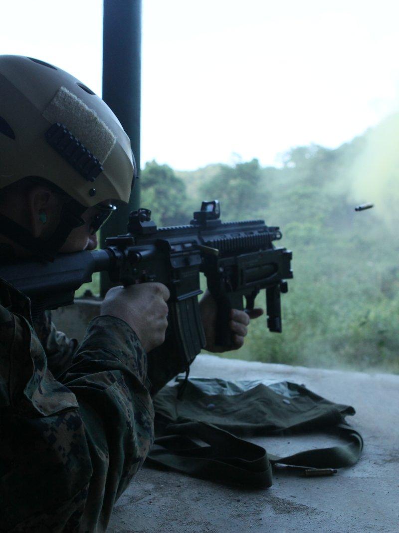 Marine in woodland camouflage and a tan helmet firing a black HK416 with a quad rail and red dot, spent cases flying toward yellow smoke on a jungle range