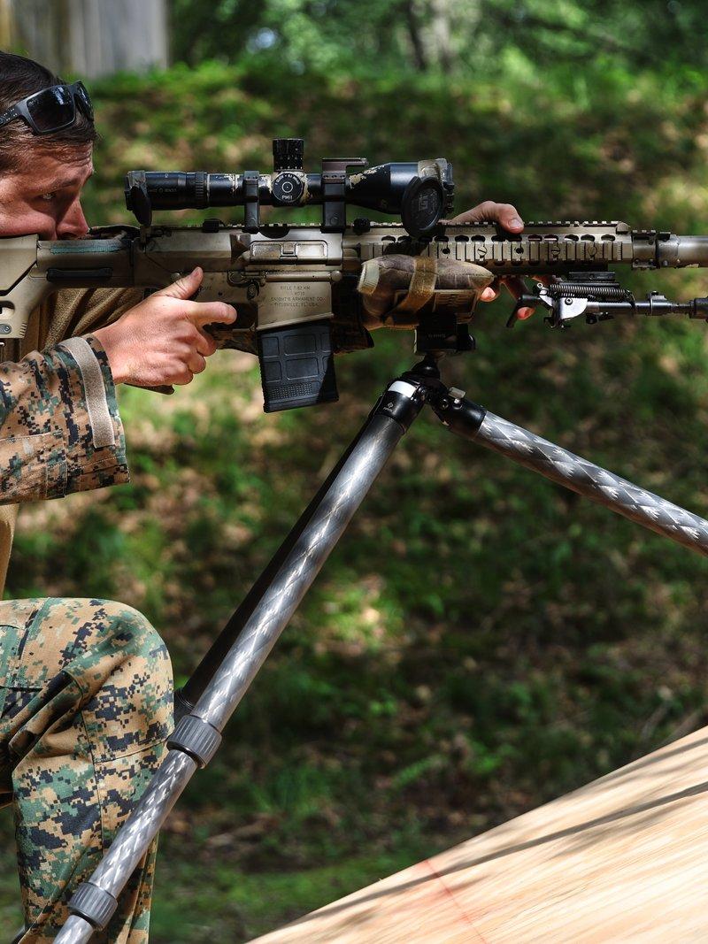 Un Marine con camuflaje boscoso arrodillado detrás de un M110 montado en un trípode, con una culata ajustable y una mira negra