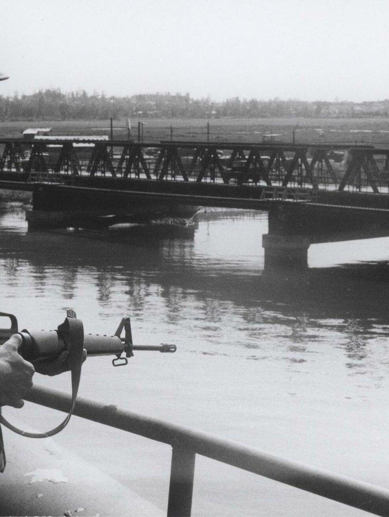 Black and white photo of a Marine in a utility cap holding a carry handle M16 with a sling, standing above a river with a steel truss bridge behind him