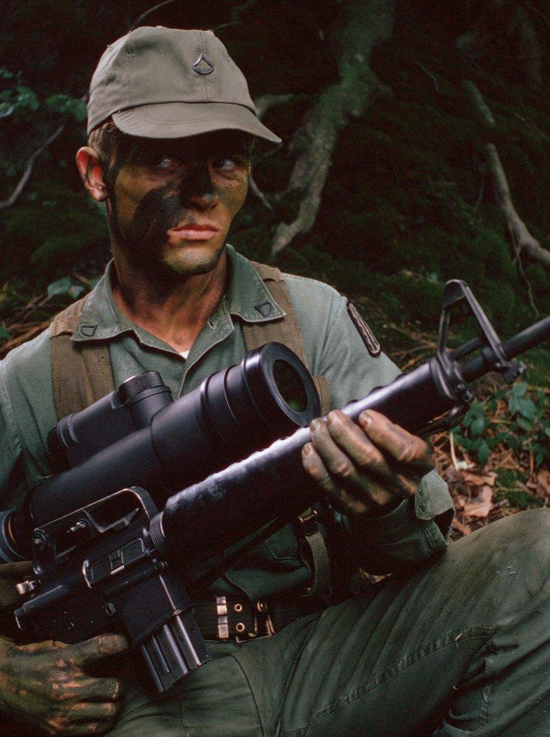 Soldier in olive green utilities and face paint sits in the woods holding an M16A1 with a large AN/PVS-2 Starlight night scope mounted on the carry handle