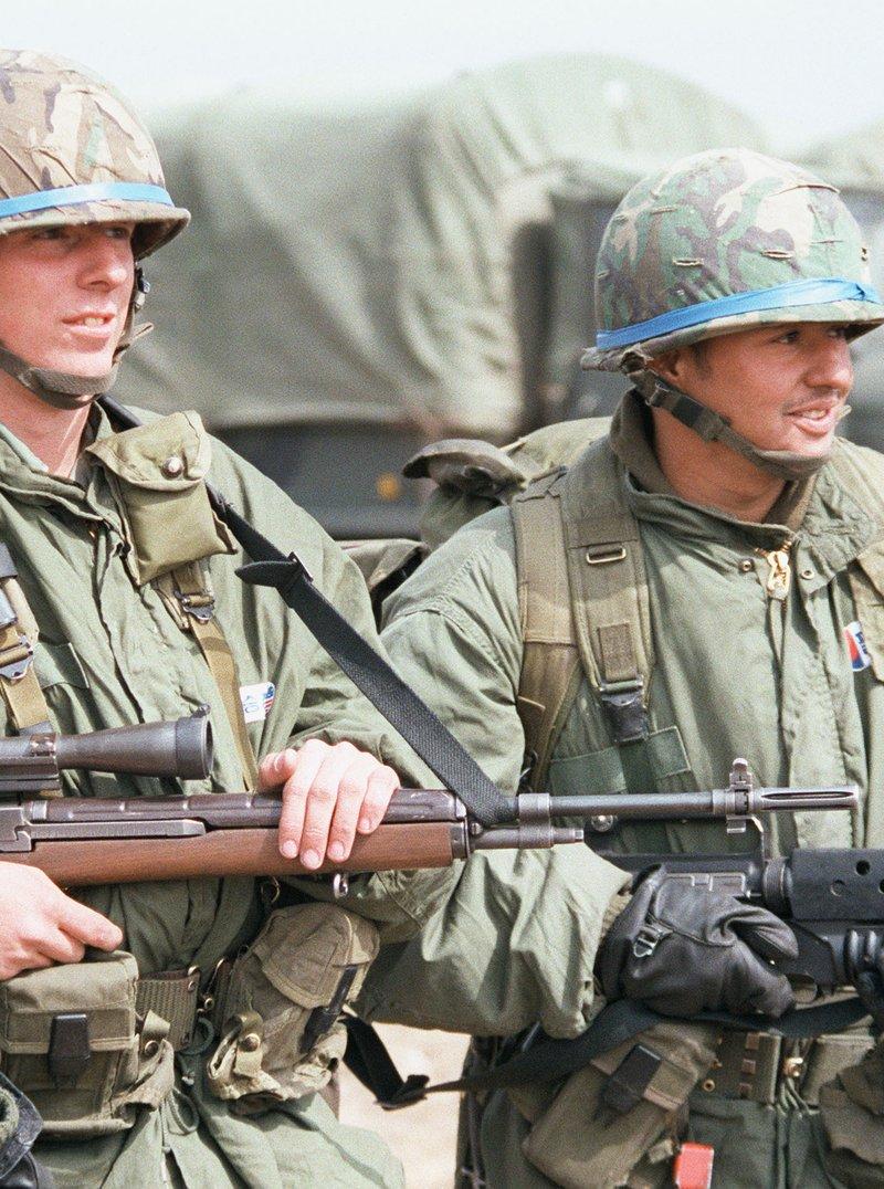 Two soldiers in M1 helmets and field jackets stand in front of army trucks, one holding a scoped M21 rifle and the other an M16A1 with an M203 grenade launcher under the barrel