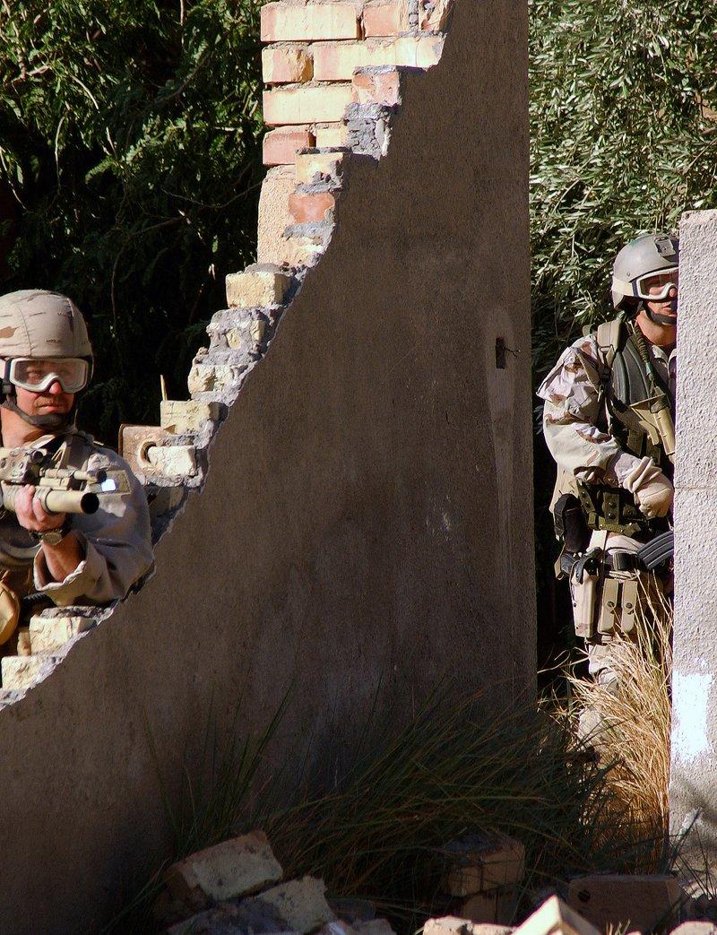 Two Air Force Pararescuemen in desert uniforms behind a broken brick wall, one aiming a tan M4 carbine with an M203 grenade launcher