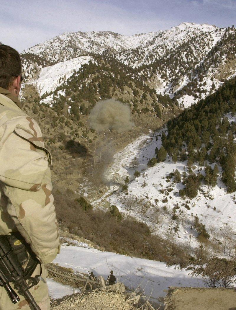 A Navy SEAL in desert camouflage with a slung M4 carbine looks across a snowy Afghan valley at a demolition blast