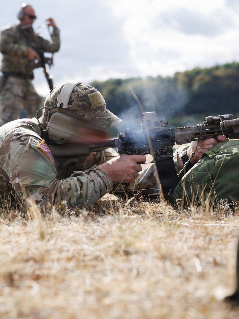 Green Beret firing prone from a suppressed M4 carbine with a tan M-LOK handguard and holographic sight on a grass range