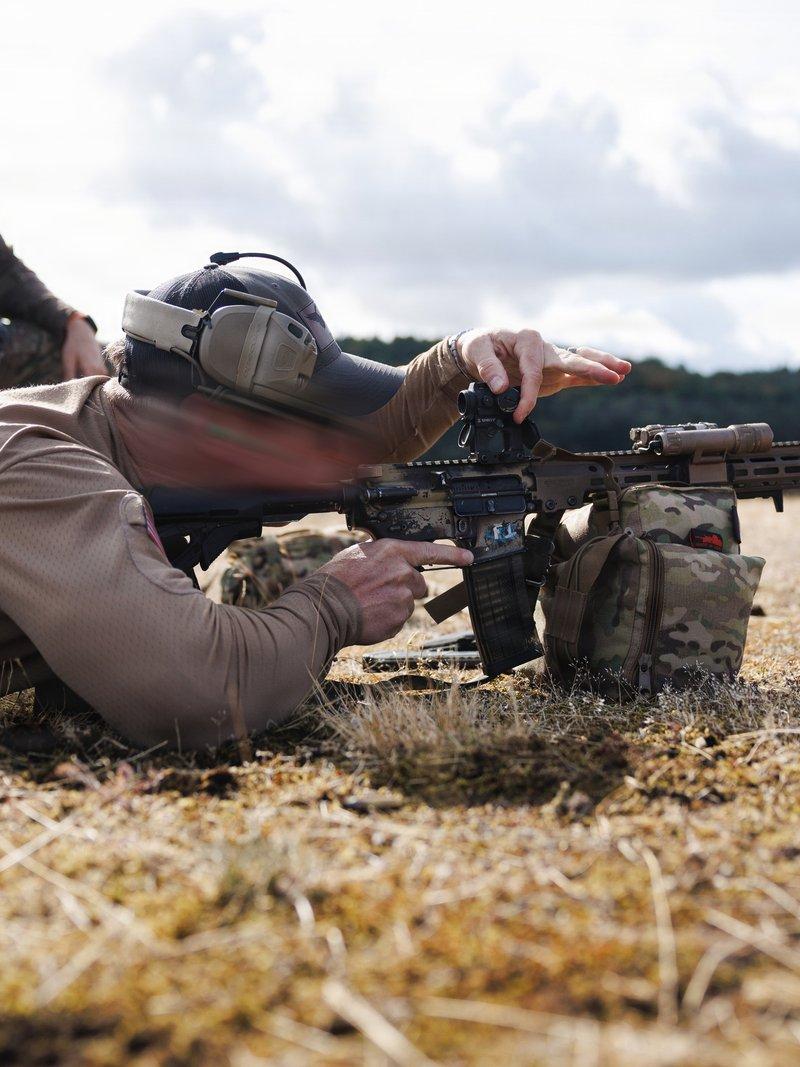 Green Beret lying prone adjusting a red dot sight on an M4 carbine with a tan M-LOK handguard, laser and weapon light