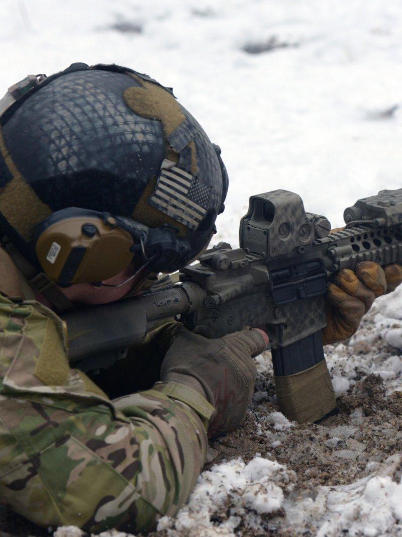 Special Forces soldier aiming a suppressed M4A1 with a quad rail handguard and holographic sight from the prone position in snow