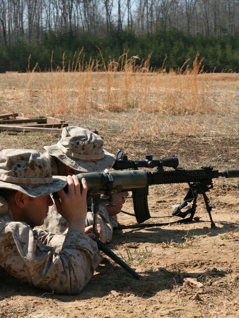 Two Marines in desert camouflage lying prone on a dirt range, one aiming a suppressed, bipod mounted Mk12 with a quad rail and scope, the other calling shots through a spotting scope