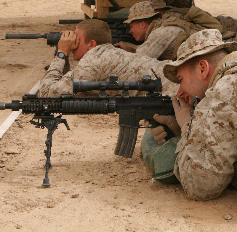 Marines lie prone on a desert range; the nearest aims a suppressed Mk12 rifle with a Knight's Armament quad rail, scope and bipod while a Marine beside him watches through binoculars