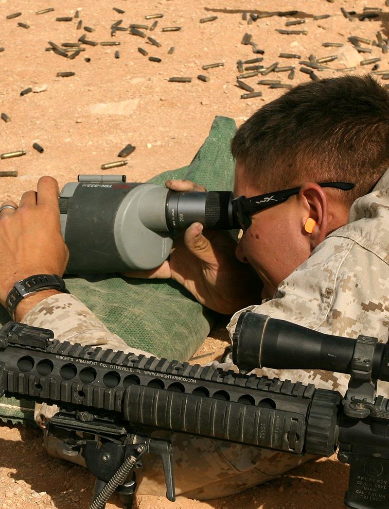 Close view of a Mk12 rifle on a range: a rail stamped Knight's Armament Co. Titusville FL, a Leupold scope in A.R.M.S. throw lever rings and a lower receiver marked M16A1, with a Marine at a spotting scope behind it