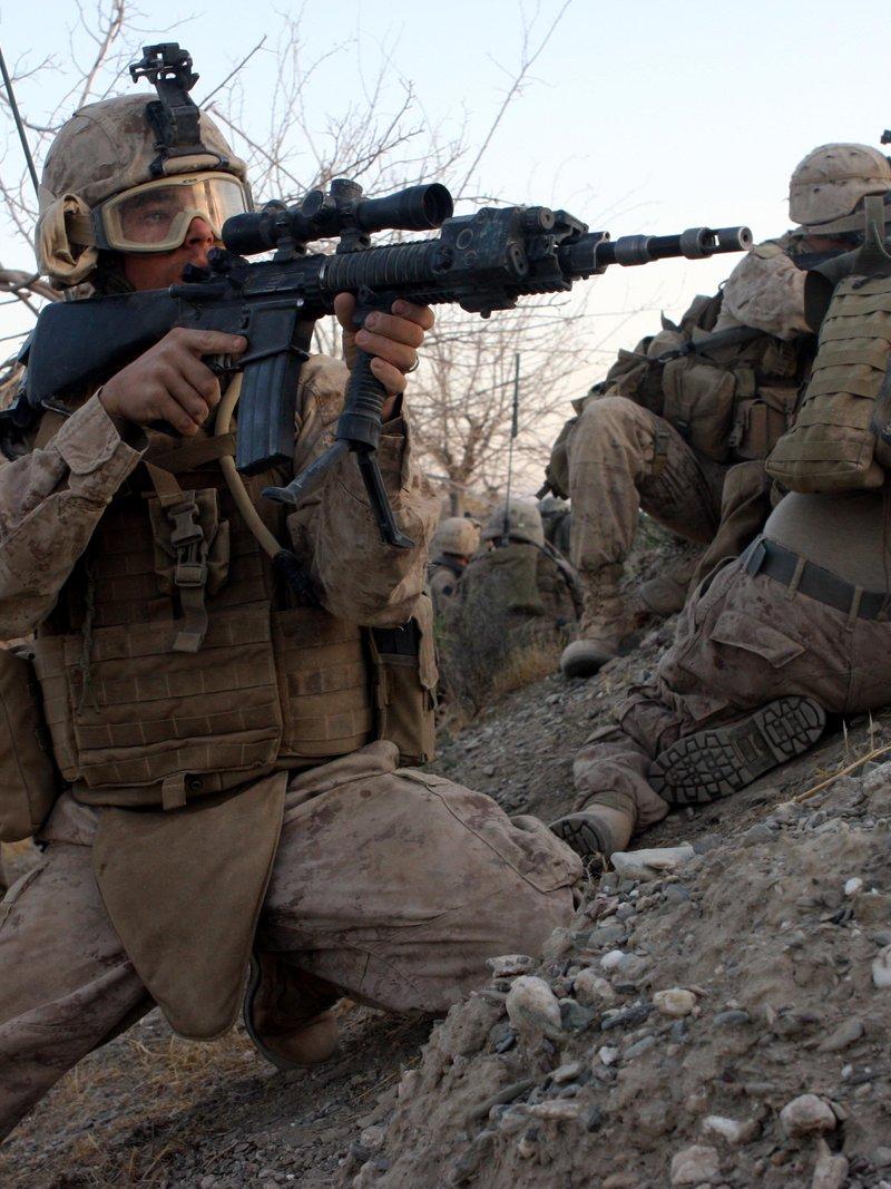 A Marine designated marksman in desert camouflage kneels at a dirt berm aiming a black rifle with a fixed stock, quad rail, laser module, scope and bipod grip, other Marines prone beside him