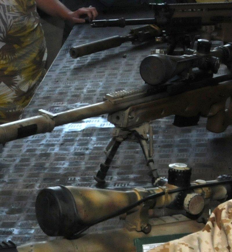 Un rifle de cerrojo pintado de camuflaje en un chasis tipo AICS sobre un bípode en una mesa de exhibición, con una mira en un riel superior largo y un supresor largo sobre el cañón, entre un FN SCAR-H y la mira de un McMillan TAC-50