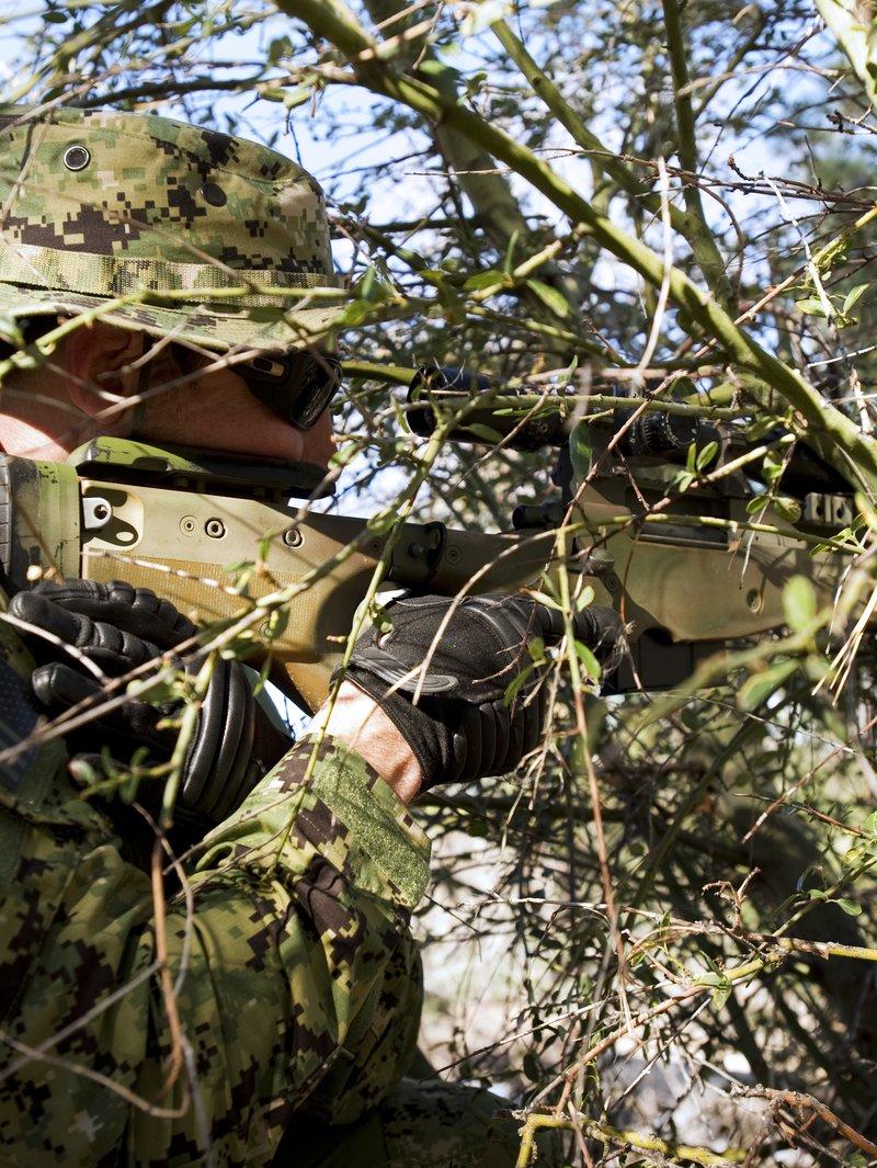 Un Navy SEAL con camuflaje digital boscoso y sombrero boonie apunta un rifle de cerrojo con mira y culata tipo AICS color tan a través de maleza espesa