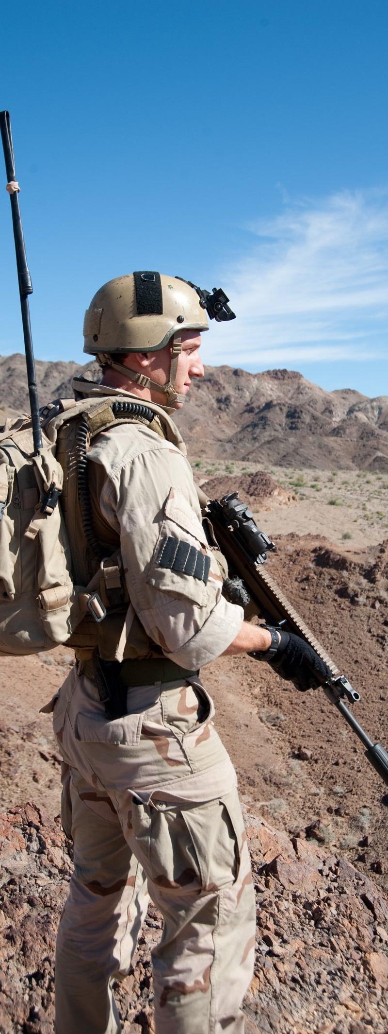Un Navy SEAL con camuflaje de desierto y una antena de radio en la mochila, de pie en una cresta rocosa sosteniendo un rifle SCAR con supresor