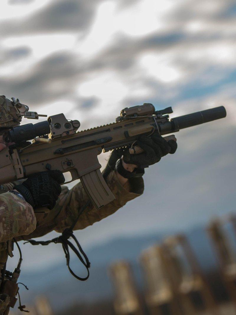 Un Ranger del Ejército de EE. UU. apunta un rifle SCAR color tan con supresor, una mira con un magnificador detrás y una unidad láser, contra un cielo nublado