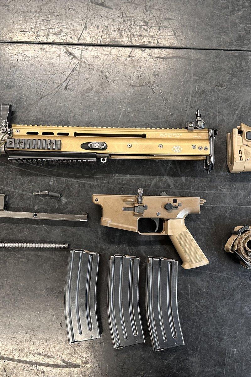 Un rifle FN SCAR-L color tan desarmado en piezas sobre una mesa de trabajo: upper receiver con cañón, culata plegable, módulo de disparador, piezas del cerrojo, resorte recuperador, tres cargadores negros y una correa tan