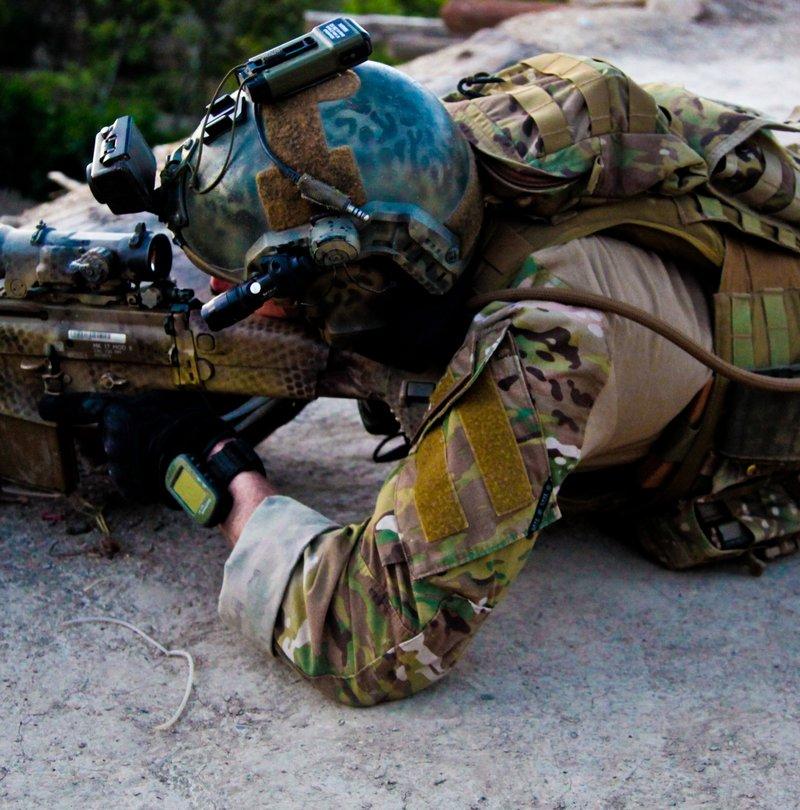 A soldier in MultiCam and a helmet with a night vision mount lies prone behind a timber beam, aiming a hand painted Mk17 with an Elcan optic and a laser module