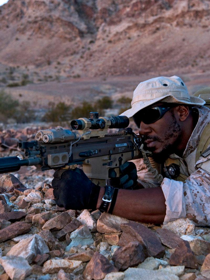 A Navy SEAL in desert camouflage and a boonie hat lies prone among rocks, aiming a suppressed flat dark earth Mk17 with a variable scope and a laser module