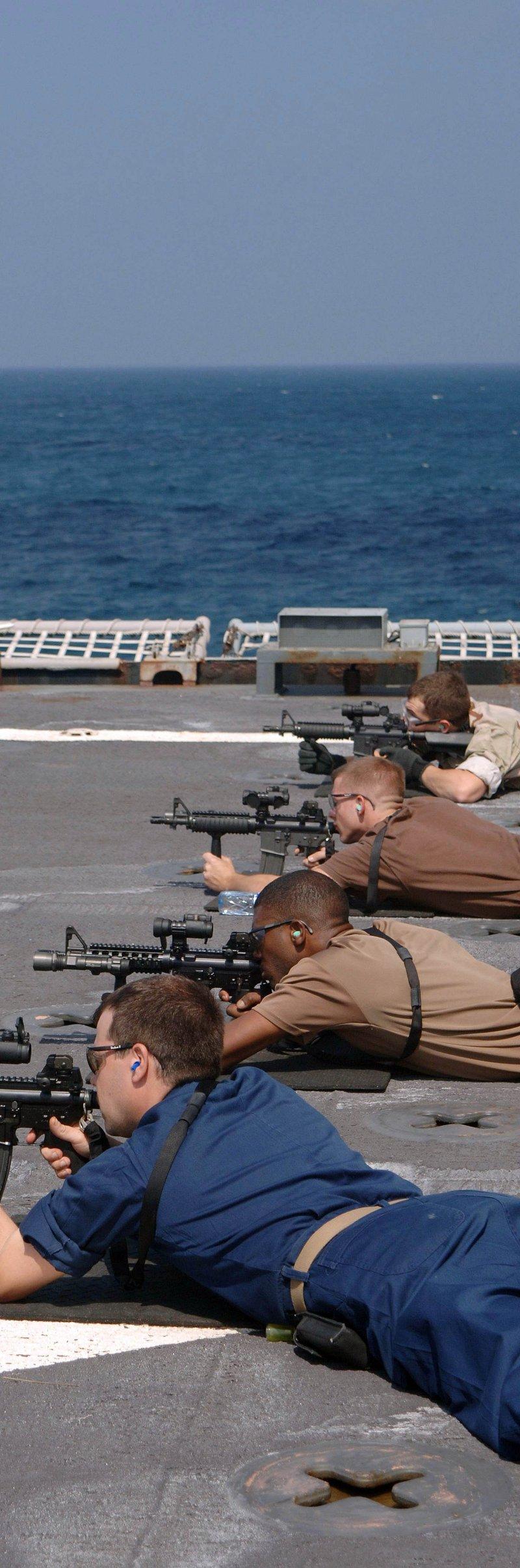 Sailors lie prone on the deck of USS Carter Hall firing MK-18 carbines with quad rails, vertical grips and red dot sights while a sailor watches through binoculars