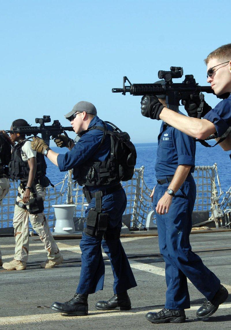 Sailors of the USS Bulkeley boarding team in blue coveralls and desert uniforms with tactical vests aim MK-18 carbines with red dot sights across the deck at sea